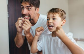 An adult and a child floss their teeth side by side in a bathroom, both wearing white t-shirts.