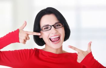 A woman with glasses, wearing a red shirt, smiles widely while pointing at her face with both hands.