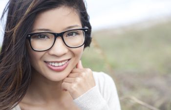 A smiling person with long hair and glasses is outdoors, with one hand resting on their chin. The background is a blurred natural setting.