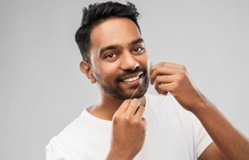 Man in a white shirt smiling while flossing his teeth against a gray background.