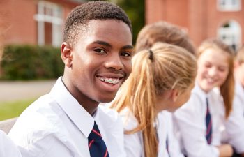 A student wearing a school uniform and braces smiles while sitting among peers outdoors, with a brick building in the background.