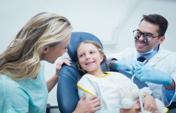 A child sits in a dental chair holding a stuffed animal, smiling at a woman beside her, while a dentist in gloves and glasses prepares to examine her teeth.