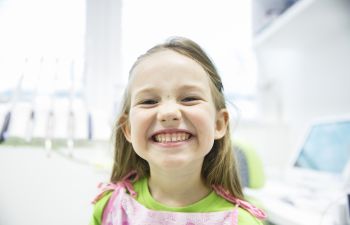 Young girl smiling widely in a brightly lit room, wearing a green top with a pink patterned overlayer.