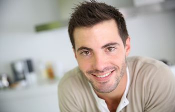 Smiling man with short dark hair and a beard, wearing a beige shirt, in a light-colored room.