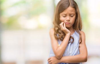 A young girl with long brown hair looks down with a thoughtful expression, resting her chin on her hand. She wears a light blue sleeveless top against a blurred outdoor background.