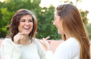 Two women in white sweaters are smiling and talking outside, with greenery in the background.