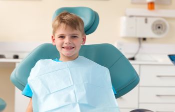 A smiling child sits in a dental chair wearing a protective bib.