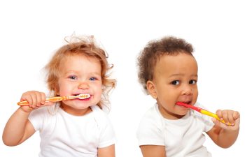 Two toddlers wearing white shirts are brushing their teeth with colorful toothbrushes against a plain white background.
