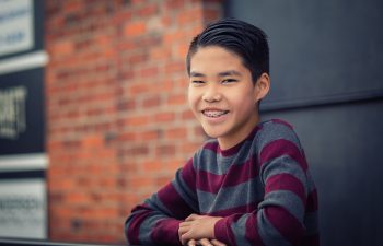 A smiling boy with short dark hair and braces leans on a railing, wearing a striped sweater in front of a blurred brick wall background.