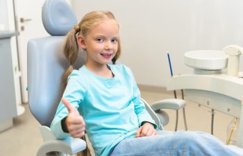 A young girl with blonde hair is sitting in a dentist's chair, smiling and giving a thumbs-up. She is wearing a light blue shirt and jeans.