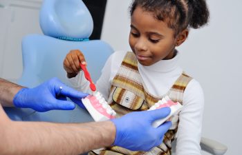 A child holds a toothbrush and learns to brush on a dental model, assisted by a person wearing blue gloves.