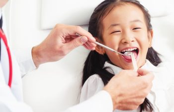 A young girl smiling while a dentist examines her teeth with a dental mirror and probe.