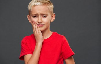 A boy with short blond hair in a red shirt holds his cheek, looking as though he might be in discomfort, against a plain gray background.