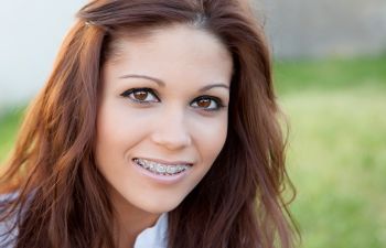 A woman with long brown hair smiles, showing braces on her teeth. She is outdoors with a blurred grassy background.