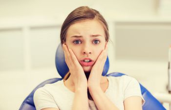 A young person sitting in a dental chair, looking surprised with hands on cheeks.