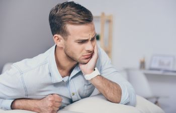 A man in a light blue shirt holds his face in apparent discomfort, sitting on a sofa in a bright room.