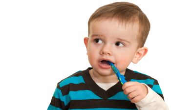 A toddler with short hair is chewing on a blue toothbrush while wearing a striped shirt, looking to the side.