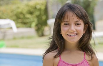Young girl smiling in a pink top, standing outdoors with a blurred green background.