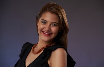 A person with long hair wears a black top and a necklace, smiling at the camera against a gray background.