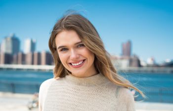 Smiling person with long hair, wearing a sweater, stands outside with a city skyline and water in the background.