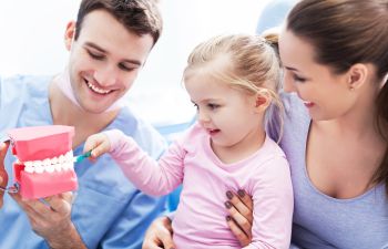 A child brushes a dental model's teeth with a toothbrush, assisted by a man and a woman.