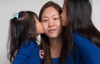A woman smiles as she receives kisses on both cheeks from two young girls, all wearing blue tops, against a plain background.
