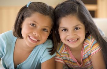 Two smiling young girls, one wearing a blue shirt and the other a striped shirt, pose closely together indoors.