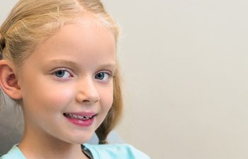 A young girl with blonde hair sits in a dental chair, smiling and holding a toothbrush. The background is a dental office.