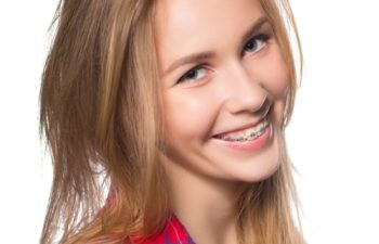 Young woman with long blonde hair wearing a red plaid shirt smiles and shows her braces.
