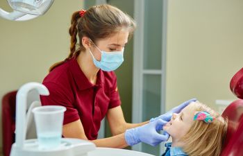 A dental hygienist wearing a mask examines a young girl's teeth in a dental office.