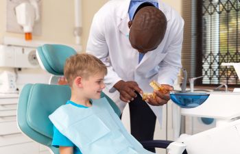 Dentist explaining dental care to a child sitting in a dental chair, using a model of teeth.