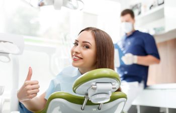 A woman sitting in a dental chair gives a thumbs-up. A dentist in the background holds a clipboard, wearing a mask and gloves.