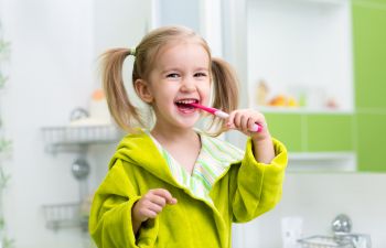 A young girl with pigtails smiles while brushing her teeth in a bathroom. She is wearing a green robe.