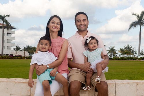A family of four seated outdoors against a backdrop of grass, palm trees, and a building, with the parents holding a young boy and girl.
