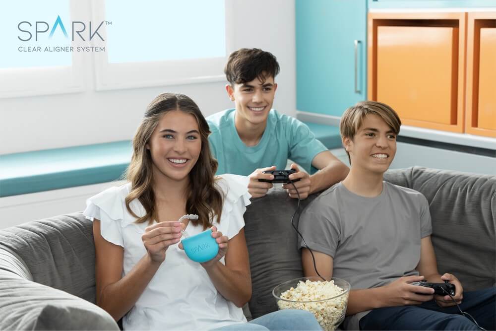 Three people sitting on a couch; two teenage boys playing video games with controllers, and a teenage girl holding a case for clear aligners. A bowl of popcorn is on the couch.