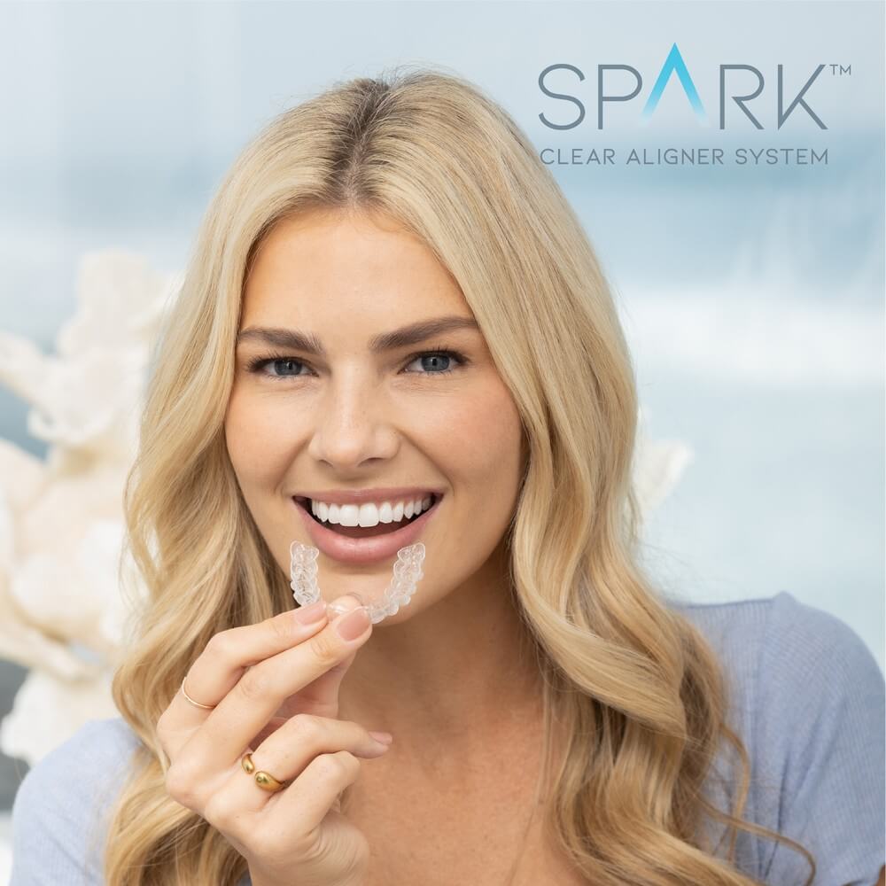 Woman smiling while holding a clear dental aligner, with "Spark Clear Aligner System" text in the background.