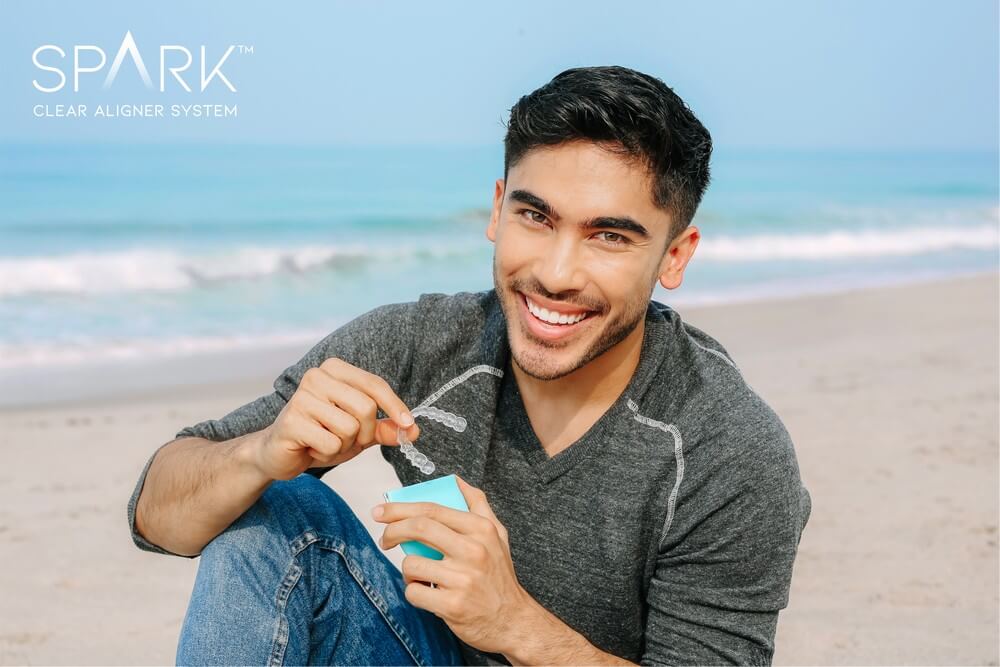 A person smiles on a beach while holding clear aligners from a box labeled "Spark Clear Aligner System.