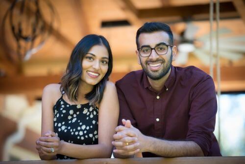 A man and woman smile while leaning on a wooden railing in a warmly lit indoor setting.