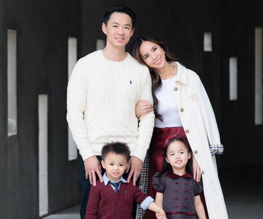 A family of four posing for a portrait in a concrete walkway. The parents stand in the back in cream and white outfits, while their two young children stand in front wearing coordinated burgundy and navy clothing.