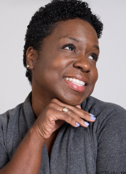 A person with short curly hair, smiling and resting their chin on their hand, wearing a gray top.
