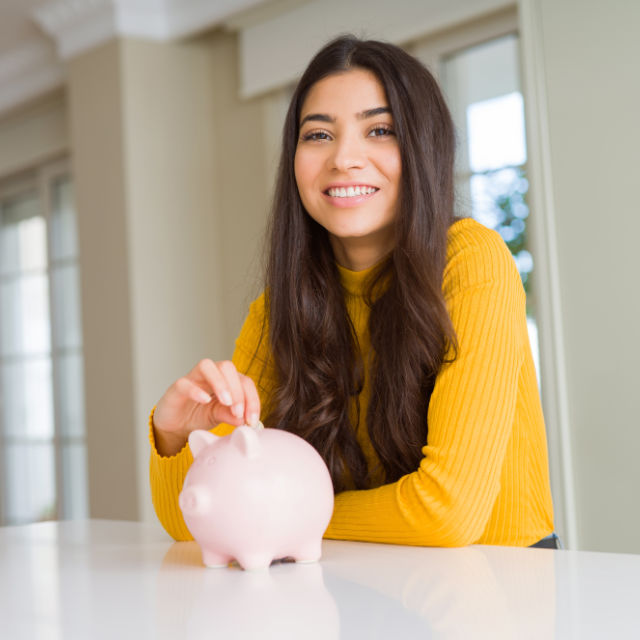 A woman in a yellow sweater places a coin into a pink piggy bank on a white table.