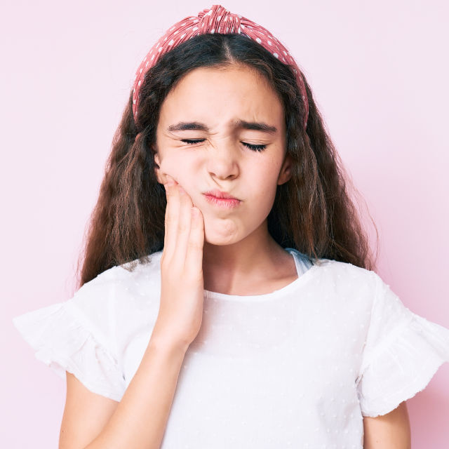 A girl with long hair and a red headband holds her cheek with a pained expression against a light pink background.