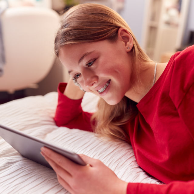 Teen girl with braces lying on a bed, wearing a red shirt, and smiling while looking at a tablet.