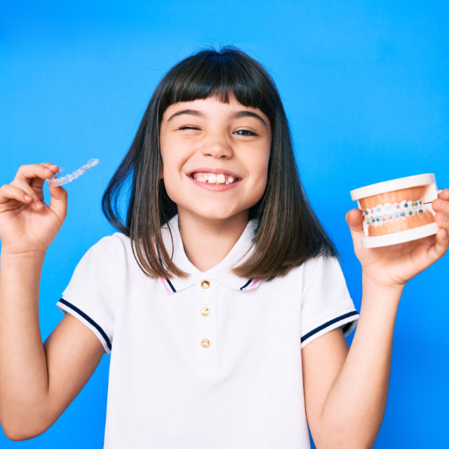A girl in a white shirt holds a dental aligner in one hand and a dental model in the other, in front of a blue background, winking and smiling.