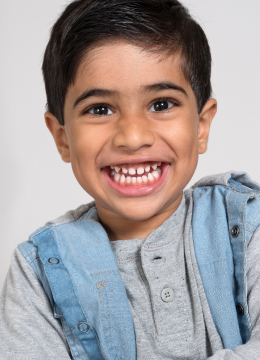 A child with short dark hair and a big smile, wearing a grey shirt and denim jacket, is facing the camera.
