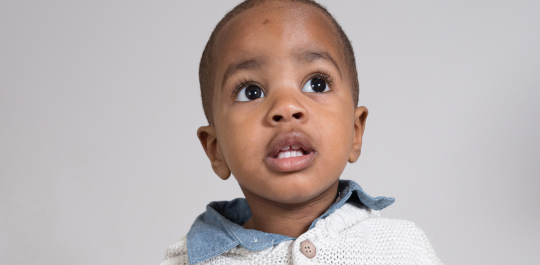 A young child with short hair looks upward, wearing a sweater over a denim shirt, against a plain background.