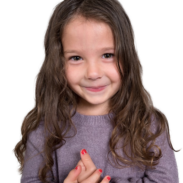 Smiling young girl with long brown hair wearing a purple sweater and red nail polish, holding her hands together.