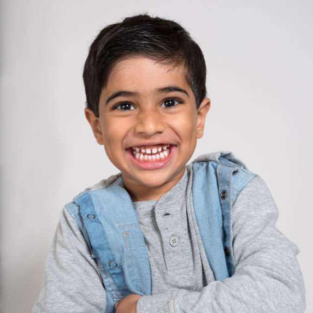 Smiling child with dark hair wearing a gray shirt and a blue denim vest, arms crossed, against a plain background.