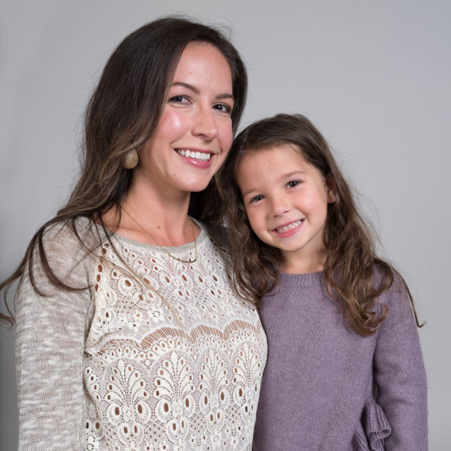 Woman and young girl standing and smiling against a gray background. The woman is wearing a lace-patterned top, and the girl is in a purple sweater.