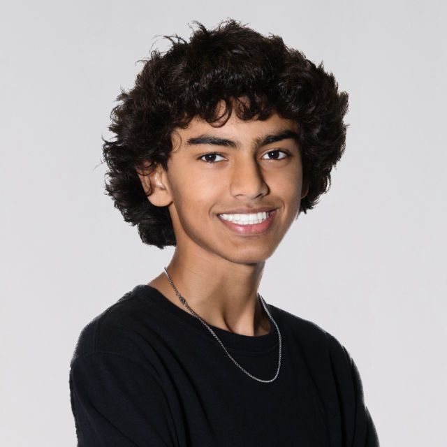A person with curly hair and wearing a black shirt and necklace smiles against a plain background.